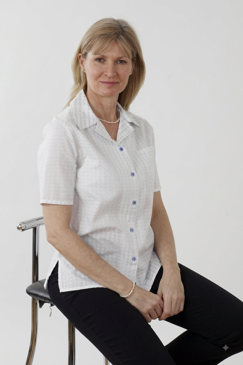 A woman with blonde hair sits on a metal chair wearing a white short-sleeve button-up shirt with a subtle blue geometric pattern and black pants, set against a plain light gray background.