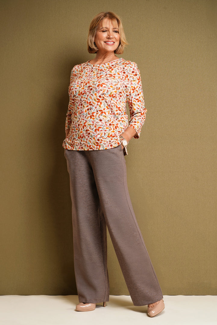 Woman wearing a Autumn leaf top with 3/4 sleeves in a warm color palette, and brown trousers,plain background.