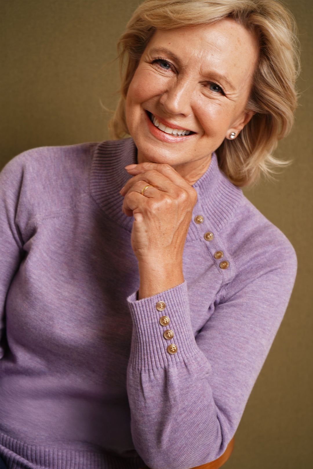Woman wearing a lilac sweater with gold buttons against a brown background