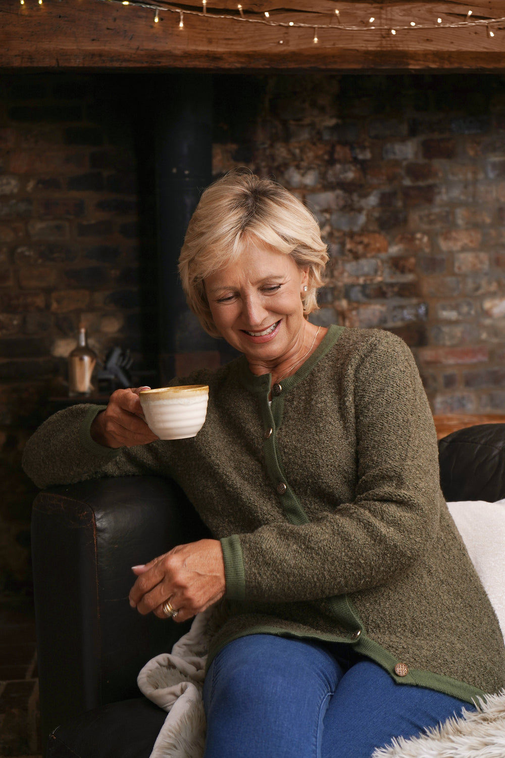 Woman sitting by a fireplace holding a cup, wearing a green cargian and blue jeans.