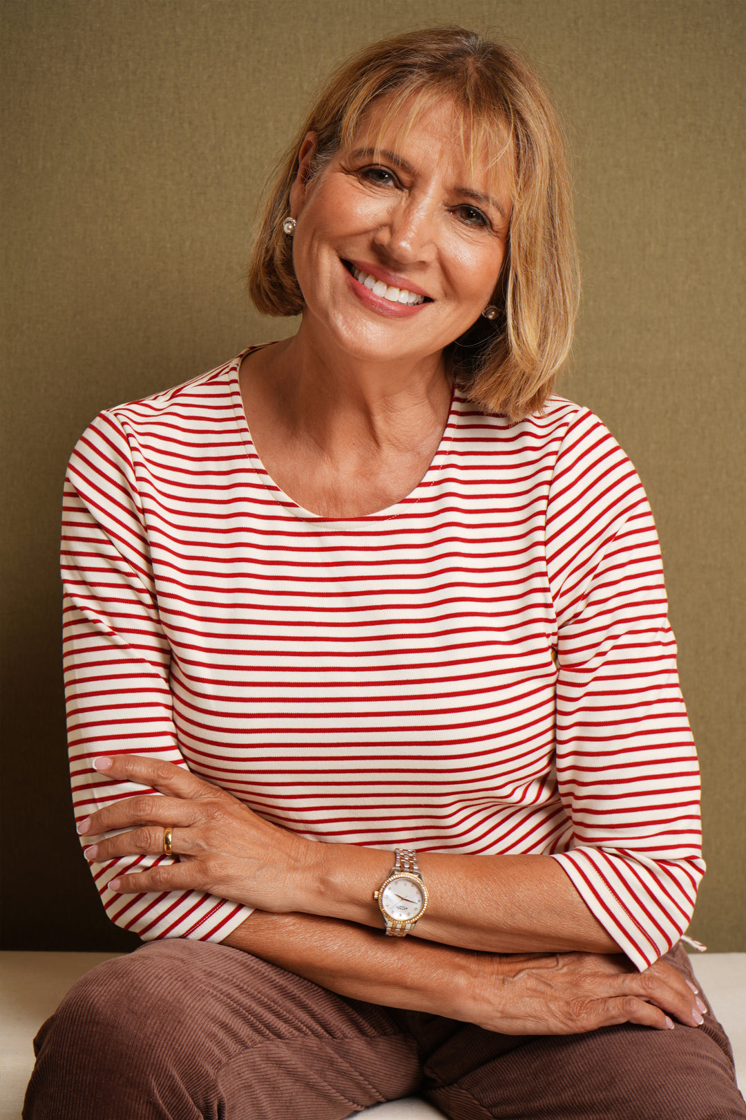 Woman wearing a red and white striped top with 3/4 sleeves and brown trousers, against a beige background.