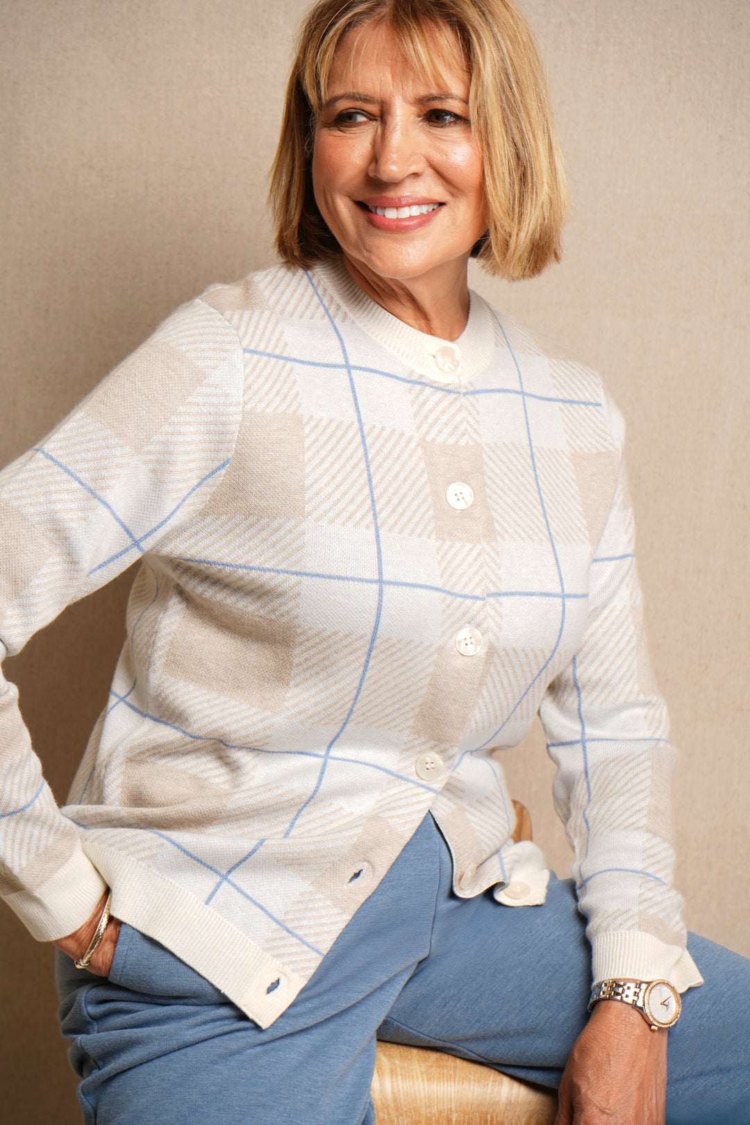 Woman wearing a white checkered cardigan and blue pants sitting against a beige wall.