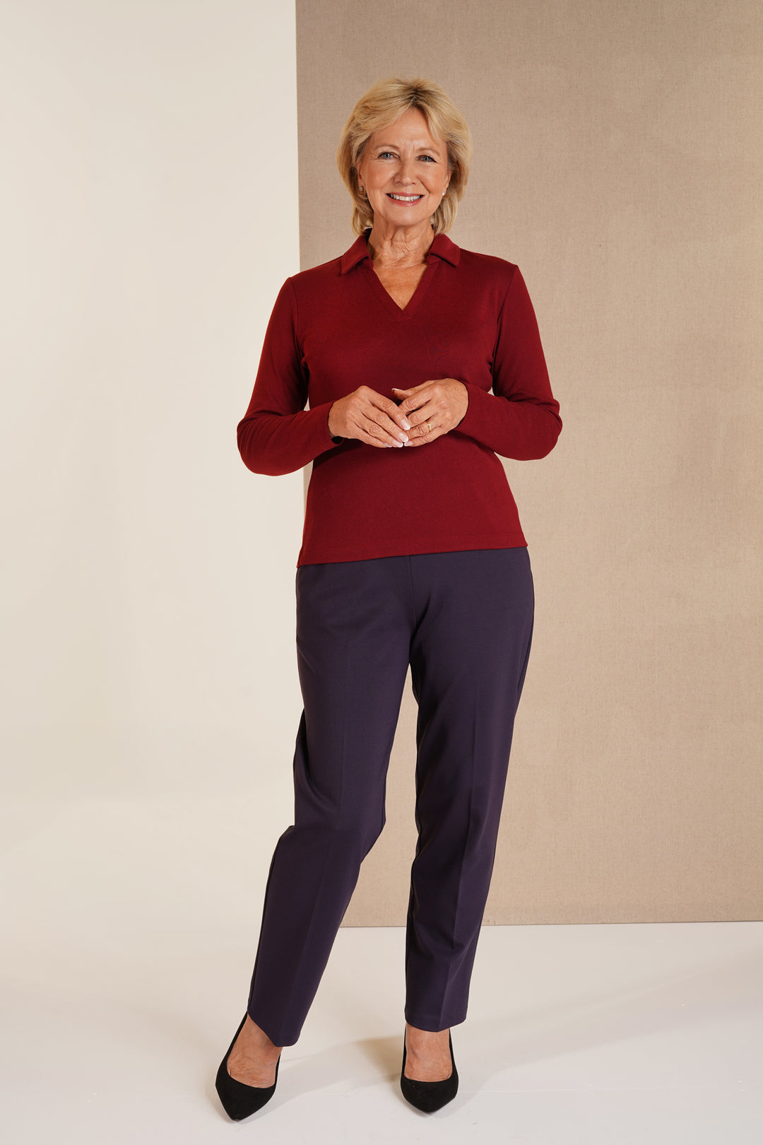 A woman modeling a long-sleeve collared knit top in deep burgundy paired with dark navy trousers and black heels, standing against a split beige and off-white studio background.