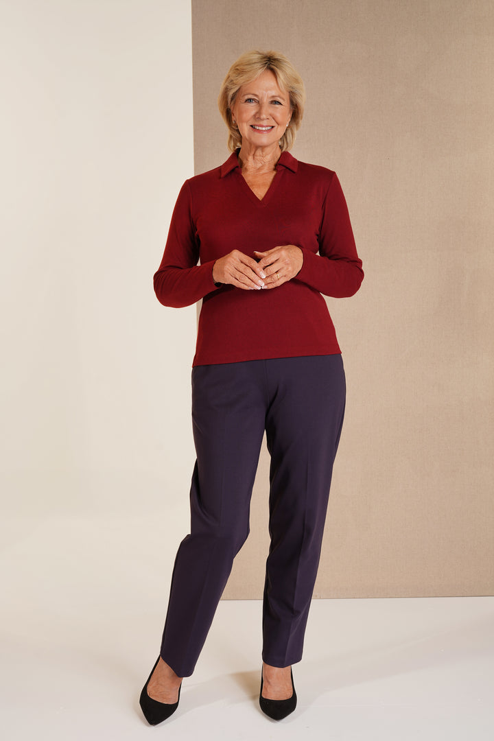 A woman modeling a long-sleeve collared knit top in deep burgundy paired with dark navy trousers and black heels, standing against a split beige and off-white studio background.