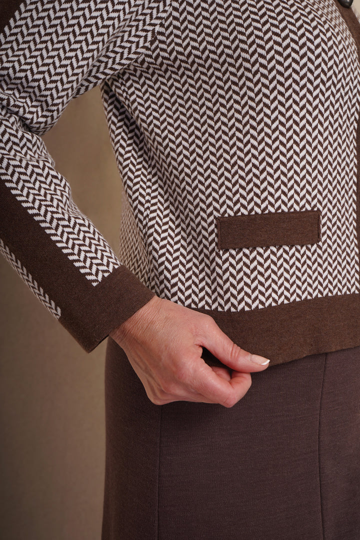 A close up of brown cardigan with a herringbone pattern, featuring mock pocket detail and dark brown trim, with brown skirt.