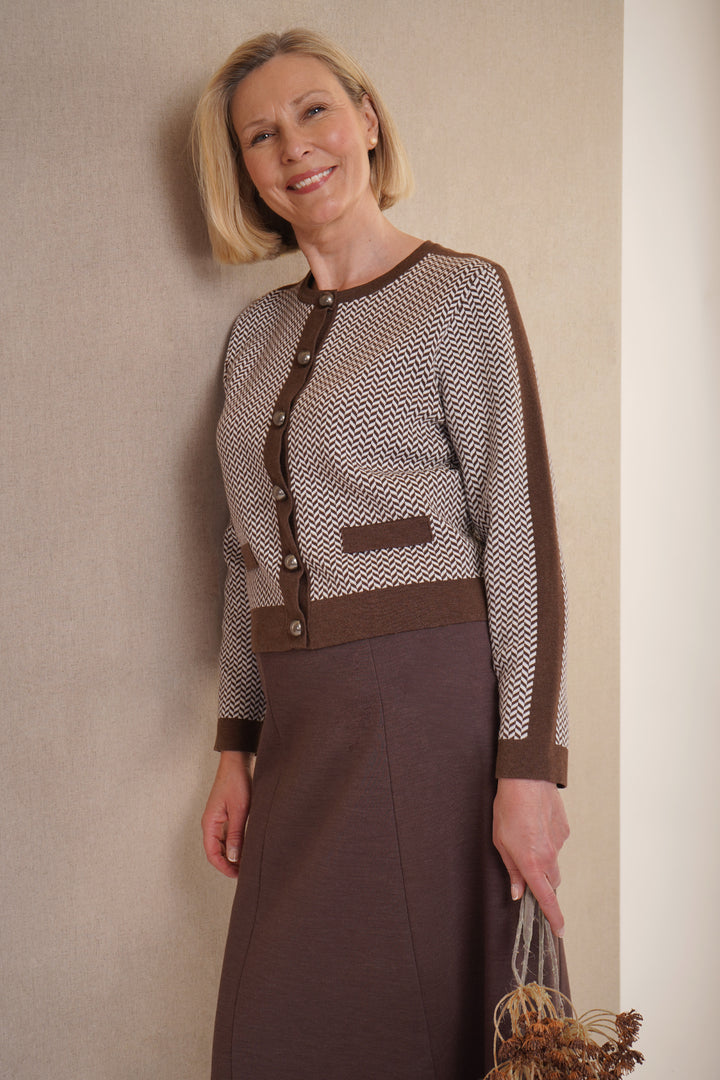 A woman standing wearing a  brown cardigan with a herringbone pattern, featuring large buttons and dark brown trim, with brown skirt.