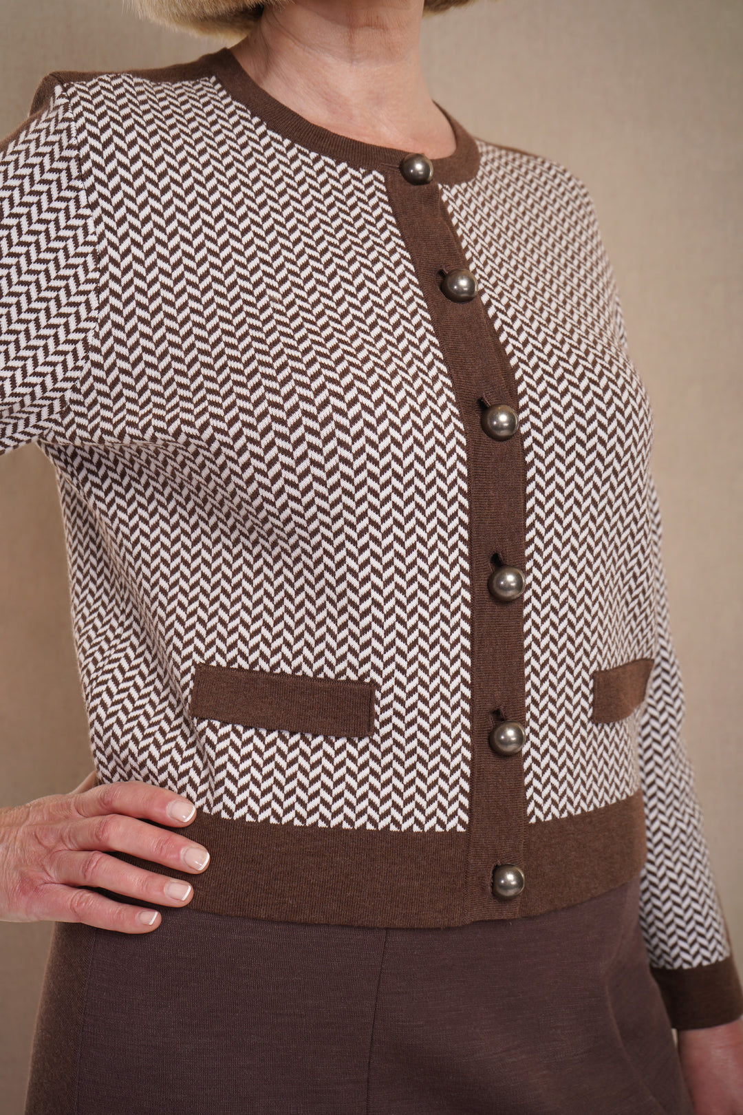 A woman wearing a  brown cardigan with a herringbone pattern, featuring large buttons and dark brown trim, with brown skirt.