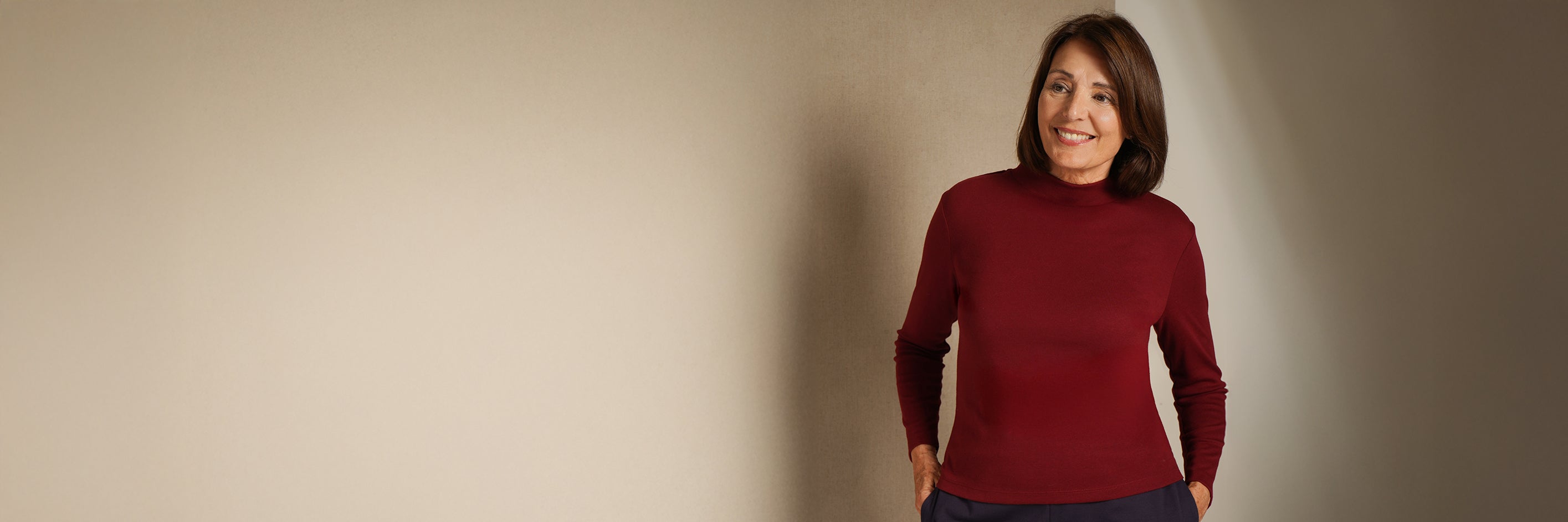 A woman with short brown hair stands against a plain beige wall wearing a fitted long-sleeve mock neck turtleneck sweater in deep burgundy red.